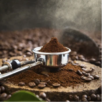Stainless steel portafilter filled with freshly ground Premium Coffee and Tea sitting on a rustic wooden block surrounded by roasted beans.Freshly ground espresso crafted from Premium Coffee and Tea beans for an authentic aroma and perfect extraction.
